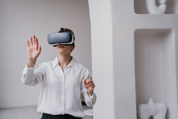 Young female student in white shirt and black pants using vr glasses exploring arts and museums standing indoors against wall with ceramics  on shelves. Girl using virtual reality. Education and fun.